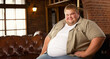 Hombre obeso y rubio, de gran tamaño, sonríe con confianza en un cómodo sofá de cuero marrón. Viste una camiseta gris y una sobrecamisa verde caqui. Su expresión muestra serenidad y satisfacción. 