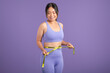 © Prostock-studio - Smiling asian woman measures waist with tape over purple studio background