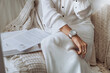 Croped woman in a white dress with a wristwatch sits on a sofa with a beige knitted blanket at home, next to her is an open book and an article, the concept of learning, self-development, reading