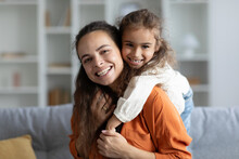 Family bonding. Portrait of happy mother and her cute daughter sharing warm embrace and smiling to camera at home, child girl hugging mom from back