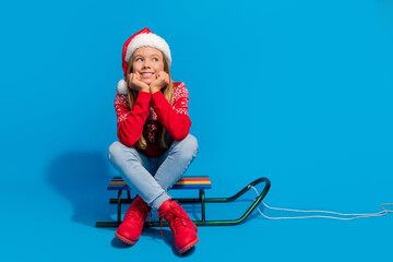 Poster - Full length photo of positive teen girl wear jumper santa hat sit on sleigh look at offer empty space isolated on blue color background