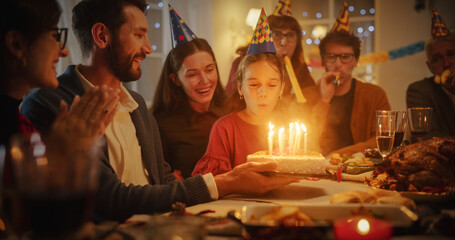 Wall Mural - Family Celebrating Birthday with a Young Girl. Father is Holding a Celebratory Cake, Little Daughter is Blowing Out Candles and Making a Joyful Wish. Happy Child is Surrounded by Her Close Relatives