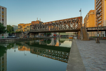 Wall Mural - Paris, France - 08 15 2021: Ourcq Canal. Reflections on the Ourcq canal of a bridge, trees, barges and buildings at sunrise.