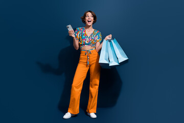 Poster - Full body photo of pretty young girl look empty space shopping bags gadget wear trendy flared pants isolated on dark blue color background