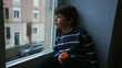 Bored sad emotion of little boy sitting by window staring at view in melancholy, child wanting to go out stuck at home