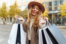 Stylish Woman In A Coat And Hat With Shopping Bags Walks Near A Shopping Center In Sunny Weather. A Beautiful Woman Enjoys The Streets Of Europe. Concept Of Consumerism, Shopping, Shopping.