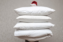 Man In Santa Hat Holding A Warm Duvet And Feather Pillows Against A Gray Wall. Stack Of Bedding For Sleeping