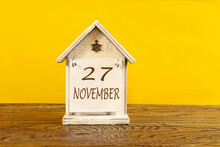 Calendar For November 27: The Name November In English, The Numbers 27 On A Decorative House On A Wooden Table, Yellow Background