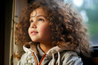 A curly-haired little girl peering out of a window with curiosity and wonder