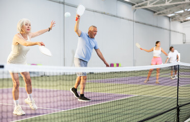 Focused positive elderly woman playing friendly pickleball match in team with male partner on indoors court. Concept of concentration in competition