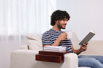 Poster - Happy man reading book and holding cup of drink on sofa with wooden armrest table at home