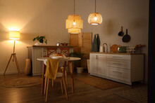 Interior of modern kitchen with glowing lamps, dining table and white counters at evening