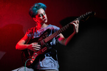Young Stylish Woman Playing Guitar Over Dark Red Background In Studio
