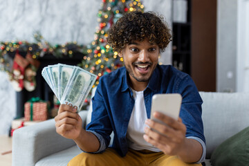 Man celebrating victory and success sitting on sofa at home in living room near Christmas tree. Hispanic man holds phone and money cash dollars in hands. A winner for the New Year holidays and