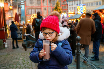 Wall Mural - Little cute preschool girl drinking hot children punch or chocolate on German Christmas market. Happy child on traditional family market in Germany, Laughing kid in colorful winter clothes