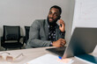 © dikushin - Portrait of Afro-American businessman in suit talking on mobile phone with client looking at laptop at camera, discussing new startup project working from office. Business man answering on phone call.