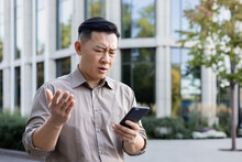Worried Senior Asian Male Office Worker Businessman Standing Outside And Using Mobile Phone. Disappointingly Looks At The Screen