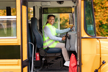 joyful black female driver sitting in yellow school bus, smiling at camera