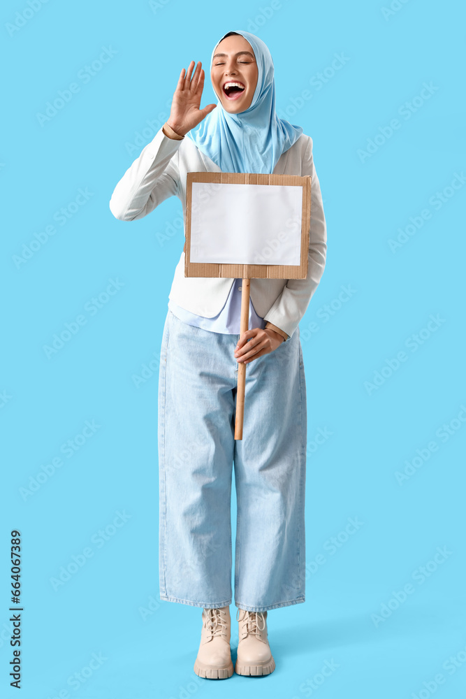 Stylish Muslim woman with blank board on blue background