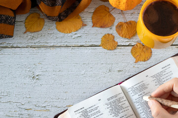 Wall Mural - Hand holding pencil over open holy bible book with a cup of coffee and autumn leaves on wooden table. Top view, copy space. Studying and reading Scriptures, Christian biblical concept.
