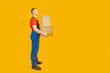 Side view portrait of full-length male loader or courier in blue jumpsuit with two boxes. Courier holds parcels on yellow background. Copy space, mock up.