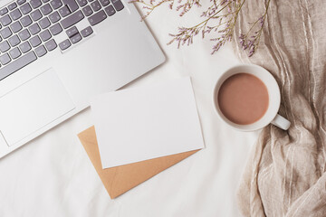 Coffee cup, laptop computer, envelope with blank card and small flowers on white linen. Cozy workplace, breakfast, comfort, hygge concept. Top view, flat lay, mockup