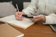 © bongkarn - Close-up image of a woman using her smartphone and taking notes in her book at her desk.