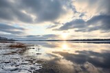 Fototapeta Niebo - Cloudy winter day at Rekyva lake with still, reflecting waters, horizon over water, and sun hiding behind white and gray clouds. Generative AI