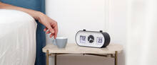 Woman In Bed Reaching For Her First Morning Coffee From The Nightstand. A Light Blue Espresso Cup On The Bedside Table Next To The Alarm Clock