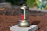 Fototapeta Krajobraz - brass grave light with red grave candle on an unplanted grave with brown soil