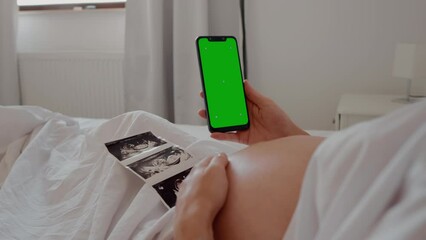Close up of pregnant woman holding smartphone with green chroma key screen and stroking tummy while sitting in bed. Technology and pregnancy concept. Future parenting.