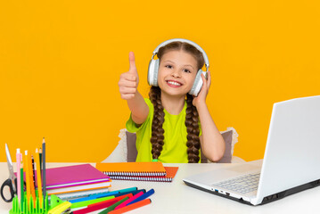 Remote online education for schoolchildren. A child with headphones is studying on a laptop. A schoolgirl does school lessons at the computer. A little girl gives a thumbs up.