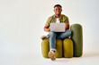 © HBS - Happy african american guy sitting on design chair and holding wireless laptop. Male freelancer in casual attire looking at camera with toothy smile over white background.