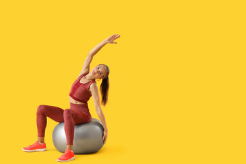 Wall Mural - Sporty young woman training with fitball on yellow background