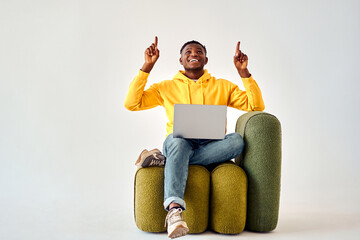 Wall Mural - Cheerful african american man in yellow hoodie and jeans sitting on green design chair with wireless laptop and pointing up on empty space. Isolated over white studio background.