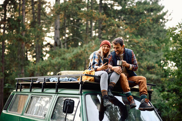 happy couple using mobile phone while sitting on roof of their camper trailer in nature.