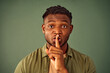 Keep my secret. Portrait of serious young man with afro hairstyle doing shush gesture near lips while standing over green background. Casually dressed black guy tying to keep conspiracy in studio.