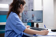 Nurse in videocall taking notes from doctor colleague in professional medical office. Woman writing down information received from general practitioner over telemedicine videoconference