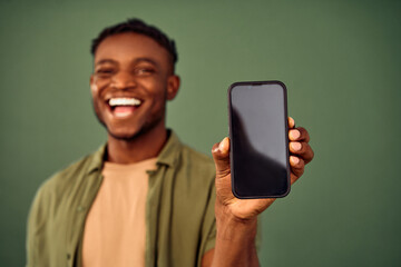 Wall Mural - App advertisement. Blurred background of joyful african american guy standing over green background and showing modern smartphone with empty black screen. Focus on cell phone with space for text.