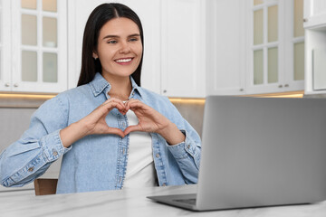 Poster - Happy young woman having video chat via laptop and making heart at table in kitchen