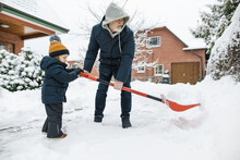 Adorable Toddler Boy Helping His Grandfather To Shovel Snow In A Backyard On Winter Day. Cute Child Wearing Warm Clothes Playing In A Snow.