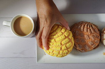 Wall Mural - Hands of Mexican Hispanic woman taking food. Concept of taking food with hands or handling food. Mexican sweet bread yellow concha on a white plate with a cup of coffee on the side 