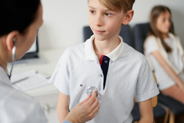 Wall Mural - Doctor and kid patient are in the clinic. Physician in white coat examining a serious young boy with a stethoscope, close up. Medicine, therapy concept