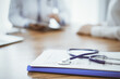 © rogerphoto - Stethoscope and medication history records form are lying on the wooden table while doctor and patient sitting and talking to each other at the background. Medicine