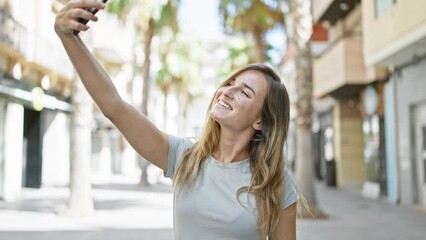 Wall Mural - Joyful young blonde woman confidently making a fun selfie with her smartphone on the city street, basking in sunlight while enjoying her urban lifestyle.