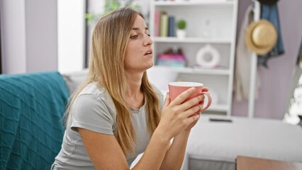 Poster - Attractive young blonde woman comfortably sitting indoors in her apartment's living room, enriching her morning by relaxing and drinking a warm espresso coffee while doubtingly pondering.