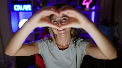 Sticker - Confident young female gamer, a radiant blonde streamer, cheerfully flashes a heart symbol with her hands during a engaging night stream in her home-based gaming room