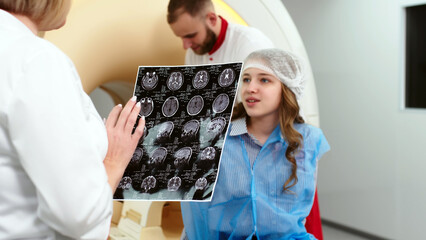 In a modern clinic, a female radiologist explains the good results of a CT scan of a young patient, observes and analyzes the CT scan next to a modern CT scanner.