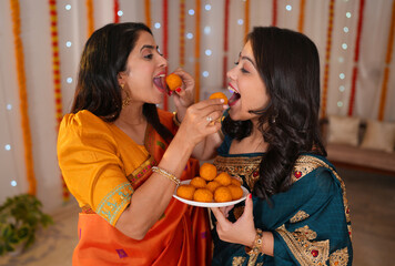 Two beautiful women celebrating the festival for Diwali at home  Decorated background. Indian sisters enjoying the festival season with sweets - Diwali celebration  sweets  decorated walls