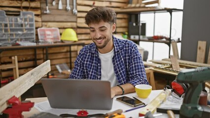 Wall Mural - Smiling young arab carpenter, basking in his well-groomed beard, engrossed in his laptop at his woodwork workshop, surrounded by timber and crafting furniture with professional gusto.